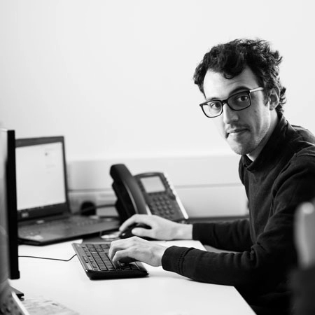 A black and white shot of Dan a Software Engineer working at a desk and looking towards the camera.