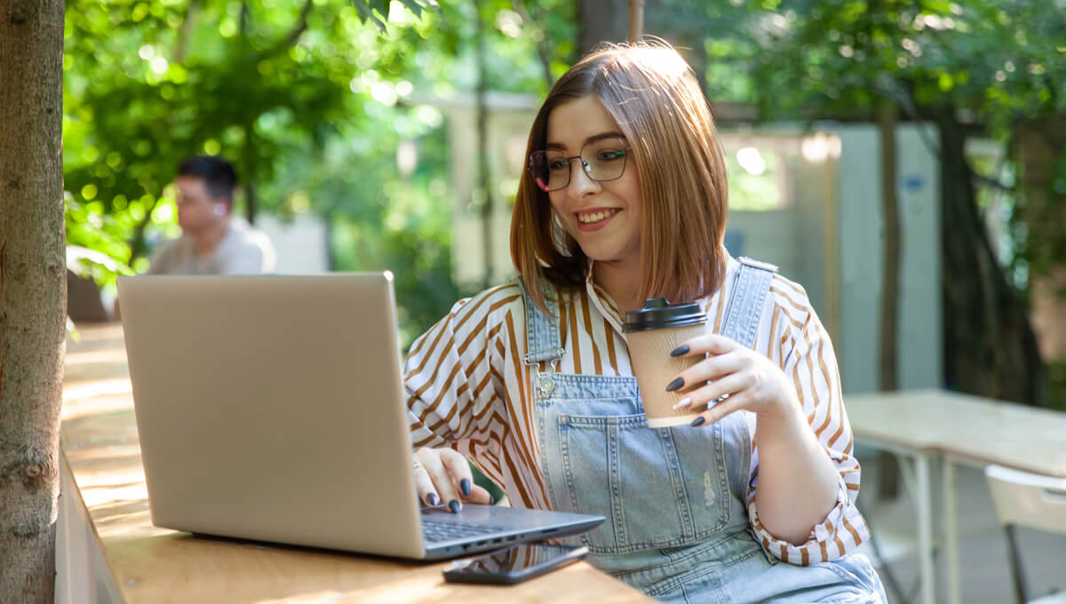 a student sitting in a cafe garden holding a coffee and working with their laptop