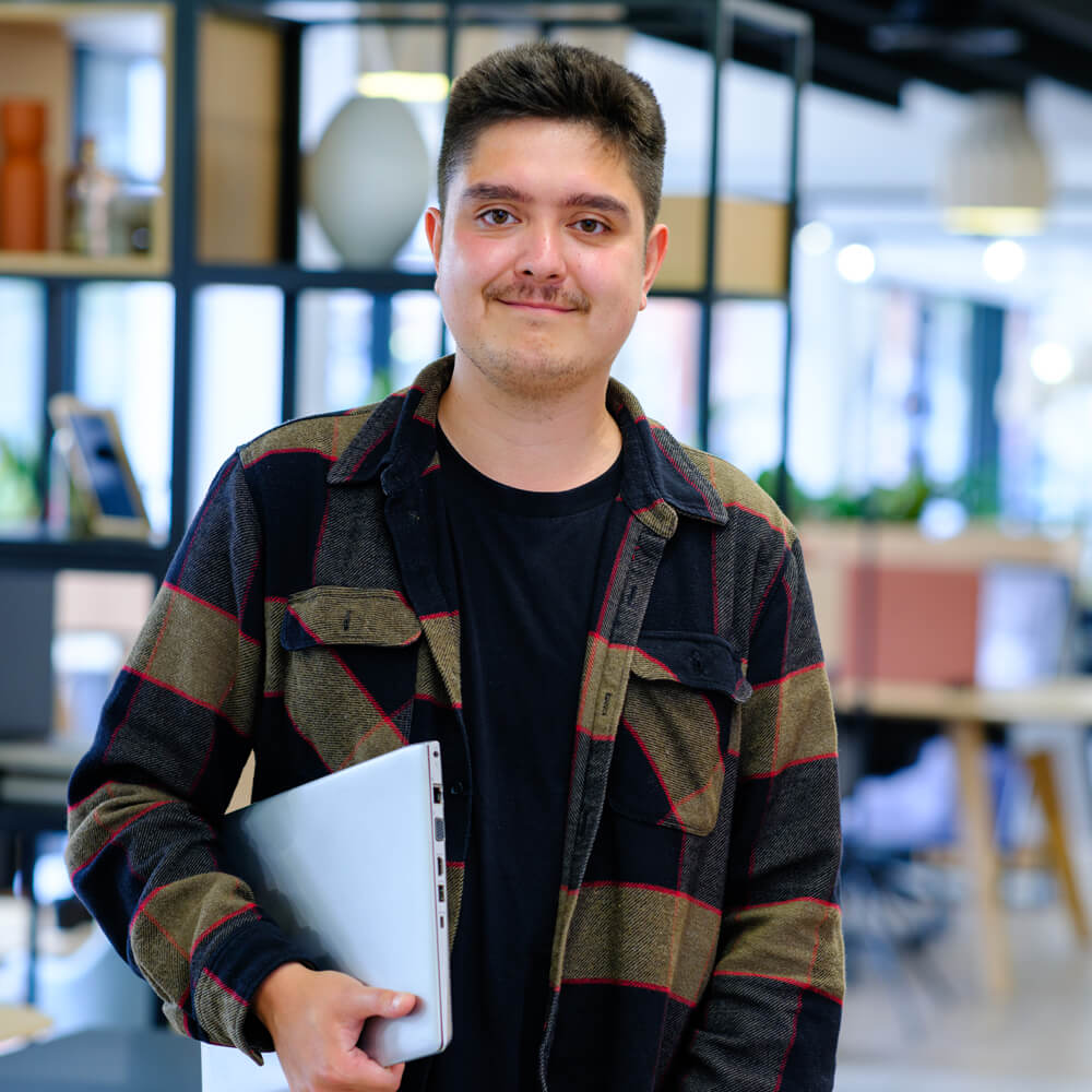 a man standing in a light office space holding a silver laptop