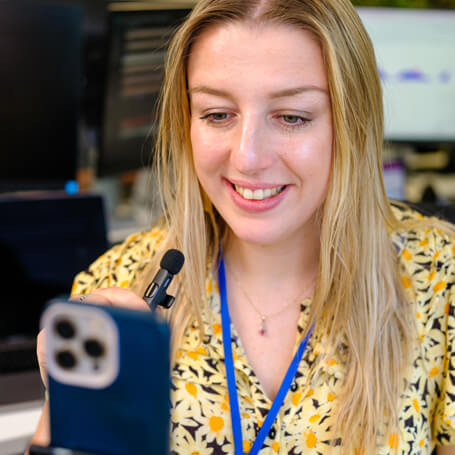 a woman smiles whilst making a social media announcement on her phone
