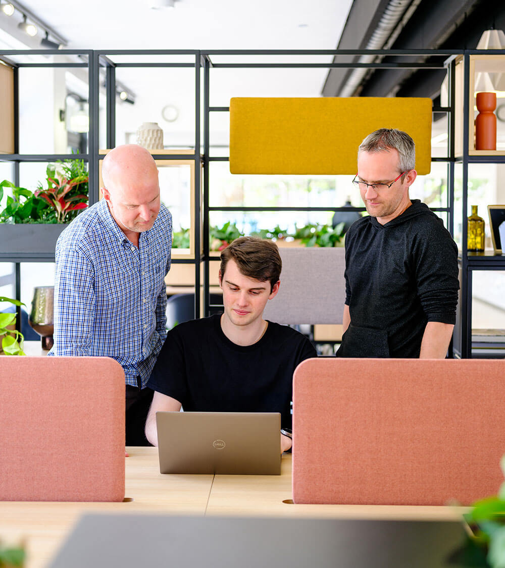 three work colleagues gather around a laptop in a bright office space